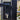 Close-up of a black Gatemaster BLD 2.0 Superlock Keyed Latch Deadlock  with plain handle; with a keyhole and a circular embossed logo on the lock plate, mounted on a gate post with an outdoor garden background visible.